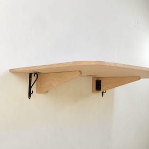 May include: A light-colored wooden wall-mounted shelf with a curved front edge. The shelf is supported by two wooden triangular brackets and two black metal hinges attached to the wall, providing a sturdy surface for storage or display.