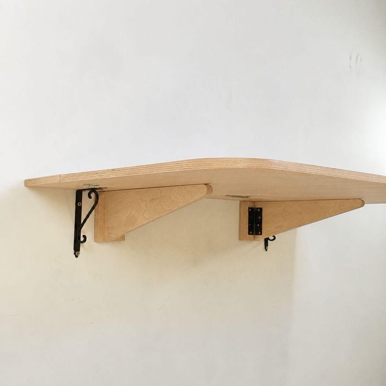 May include: A light-colored wooden wall-mounted shelf with a curved front edge. The shelf is supported by two wooden triangular brackets and two black metal hinges attached to the wall, providing a sturdy surface for storage or display.