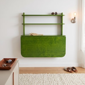 May include: A wall-mounted green wooden desk featuring a fold-down surface and two open shelves above. The desk has a visible wood grain finish and is mounted against a plain wall. Two small pinecones sit on the top shelf, and wheat stalks rest on the desk.