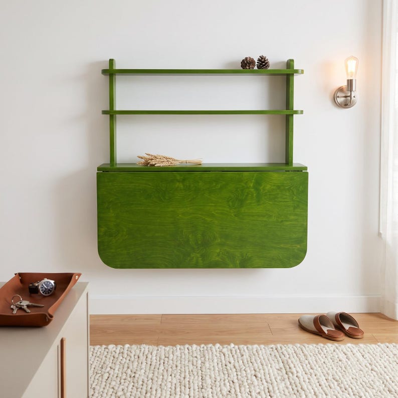 May include: A wall-mounted green wooden desk featuring a fold-down surface and two open shelves above. The desk has a visible wood grain finish and is mounted against a plain wall. Two small pinecones sit on the top shelf, and wheat stalks rest on the desk.