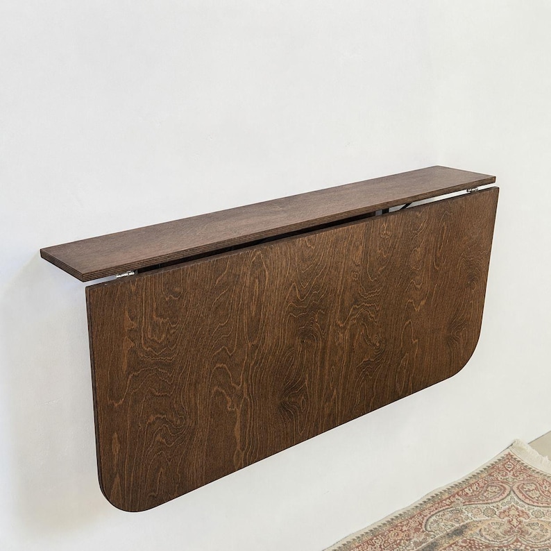 May include: A wall-mounted, dark brown wooden drop-leaf table featuring a rectangular top shelf and a large, hinged, semi-circular panel that folds downward. The wood grain is visible across the surface, and the structure is attached to a wall.