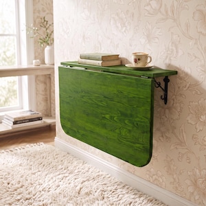 May include: A wall-mounted, fold-down table featuring a vibrant green wood grain finish. The table is attached to the wall with black metal brackets and supports two stacked books and a ceramic coffee cup with a floral pattern on its surface.