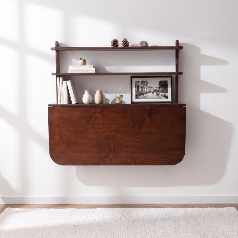 May include: A wall-mounted dark wood drop-leaf desk featuring two upper shelves. The shelves hold books, small decorative objects, and a framed black and white photograph. The desk surface is folded down, displaying a smooth, rich wood grain finish.