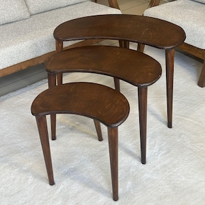 Puede incluir: Un juego de tres mesas nido hechas de madera de color marrón oscuro. Cada mesa presenta una superficie superior lisa en forma de riñón y tres patas cónicas. Las mesas están dispuestas en formación de cascada, con la más grande atrás y la más pequeña al frente, mostrando un acabado de veta de madera consistente en las tres piezas.