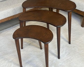 Puede incluir: Un juego de tres mesas nido hechas de madera de color marrón oscuro. Cada mesa presenta una superficie superior lisa en forma de riñón y tres patas cónicas. Las mesas están dispuestas en formación de cascada, con la más grande atrás y la más pequeña al frente, mostrando un acabado de veta de madera consistente en las tres piezas.