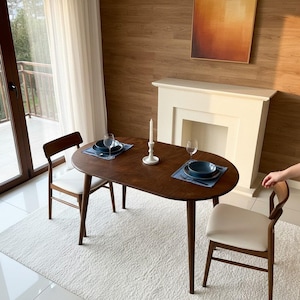 May include: An oval dark wood dining table with four tapered legs, accompanied by two chairs featuring dark wood frames and white upholstered seats. The table is set with two blue plates, two clear wine glasses, and a white candle holder.