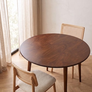 May include: A round dark brown wooden dining table with a smooth surface and four tapered legs, accompanied by two chairs featuring light wood frames, woven cane backrests, and textured off-white upholstered seats arranged around the table.