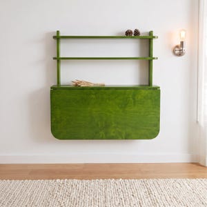 May include: A wall-mounted, olive green wooden desk featuring a fold-down surface and two open shelves above. The desk has a smooth, stained wood grain finish and rounded corners on the drop-leaf panel, with two small pinecones on the top shelf.