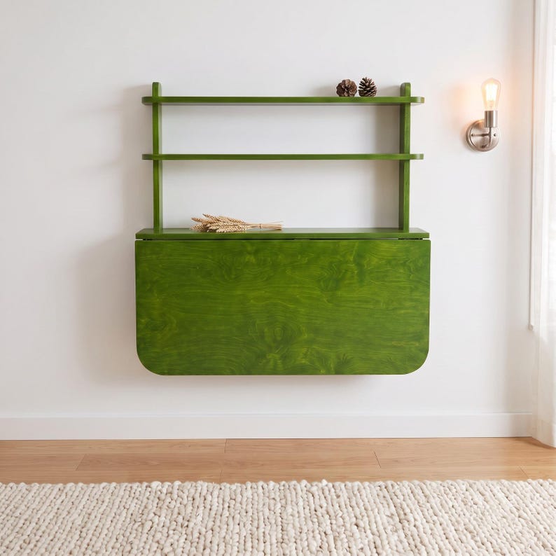 May include: A wall-mounted, olive green wooden desk featuring a fold-down surface and two open shelves above. The desk has a smooth, stained wood grain finish and rounded corners on the drop-leaf panel, with two small pinecones on the top shelf.