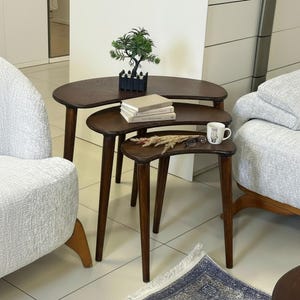 Puede incluir: Un juego de tres mesas nido de color marrón oscuro, con forma de riñón y patas cónicas. Las mesas están dispuestas en forma escalonada, con un pequeño árbol decorativo, libros, una taza y gafas encima. Muebles claros alrededor.