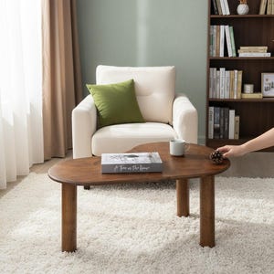 Könnte beinhalten: Ein brauner Couchtisch aus Holz mit einer einzigartigen, organischen Form. Der Tisch hat vier zylindrische Beine. Ein Buch, eine Tasse und ein Tannenzapfen liegen auf dem Tisch. Der Tisch steht vor einem weißen Sessel und einem Bücherregal.