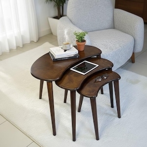 Puede incluir: Juego de tres mesas nido de color marrón oscuro con bordes curvos y patas delgadas y cónicas. Una pequeña planta en maceta, libros, una tableta y gafas descansan sobre las mesas. Las mesas están sobre una alfombra blanca, cerca de un sillón gris.