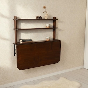 May include: A dark brown wall-mounted folding desk featuring two upper shelves. The desk surface is in a vertical folded position. The shelves hold two small pinecones, a white vase with a dried flower, two ceramic mugs, and two stacked books.