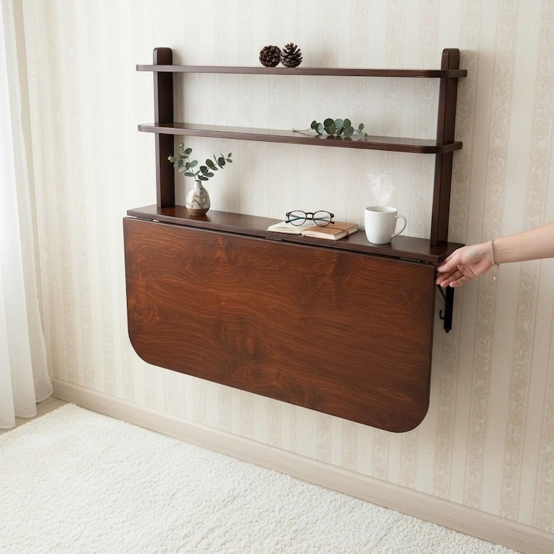 May include: A dark brown wooden wall-mounted desk featuring a fold-down tabletop and two upper shelves. The unit includes a flat surface for items, with a white mug, black-rimmed glasses, and a small notebook placed on the top shelf area.