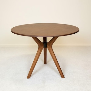 May include: A round dining table crafted from dark brown wood with a smooth, circular tabletop surface. The table features three angled wooden legs that converge at a central black cylindrical connector piece, providing a stable base structure.