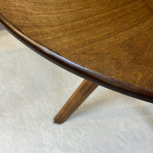 May include: A round wooden table with a smooth, medium-brown stained surface and a visible wood grain pattern. The table features a single tapered leg extending downward from the underside, constructed from the same dark-toned wood material.