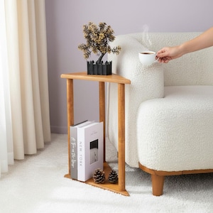 Puede incluir: Una mesa auxiliar triangular de madera con un estante inferior. La mesa contiene dos libros, dos piñas y una pequeña maceta negra con una planta decorativa. Una persona sostiene una taza de té blanca cerca de un sofá blanco.