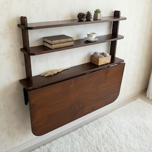 May include: A dark brown wall-mounted wooden unit featuring two upper shelves and a large, fold-down drop-leaf table. The shelves hold two books, a white teacup on a saucer, two pinecones, a small potted succulent, and a woven basket with yarn.