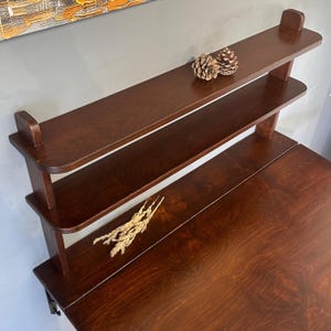 May include: A dark brown wooden wall-mounted desk featuring two upper shelves with rounded edges. The desk surface is flat and rectangular, while the shelves are supported by vertical wooden brackets. Two pinecones sit on the top shelf.