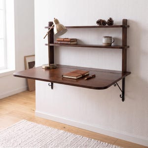 May include: A wall-mounted dark wood desk featuring a fold-down work surface supported by metal brackets, two upper shelves, a brass-toned desk lamp, two stacked leather-bound books, a single notebook, a pen, a ceramic mug, and two pinecones.
