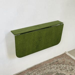 May include: A wall-mounted, olive green wooden drop-leaf table in a folded position. The table features a rectangular top shelf and a larger, rounded-corner panel that hangs vertically against the wall, showcasing a visible wood grain texture.