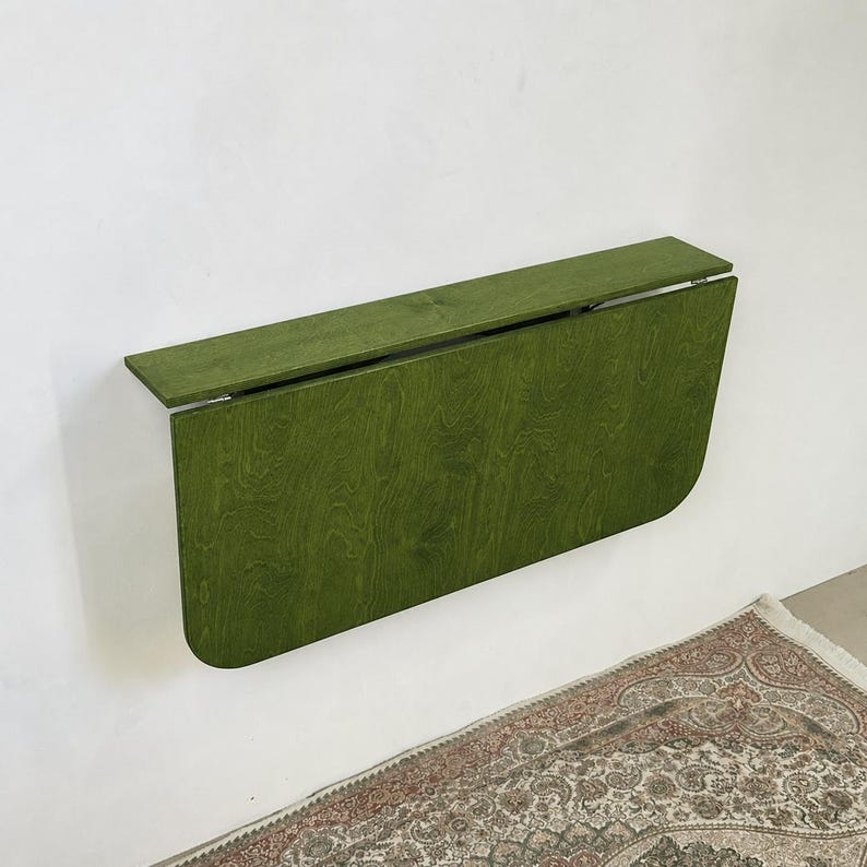 May include: A wall-mounted, olive green wooden drop-leaf table in a folded position. The table features a rectangular top shelf and a larger, rounded-corner panel that hangs vertically against the wall, showcasing a visible wood grain texture.