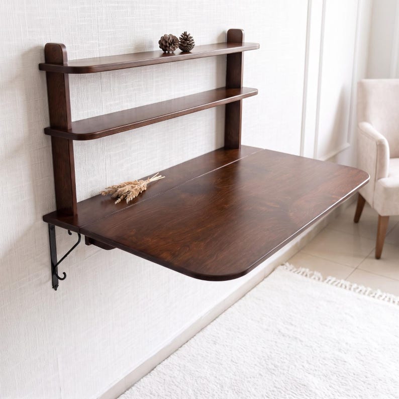May include: A wall-mounted dark brown wooden desk featuring a smooth, rectangular surface with rounded corners and two integrated upper shelves. The desk is supported by black metal brackets and includes two small pinecones and dried wheat stalks.