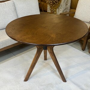 May include: A round wooden table with a dark brown finish and a visible wood grain pattern. The table features three angled legs that meet at a central point beneath the circular top, providing a stable base for the furniture piece.