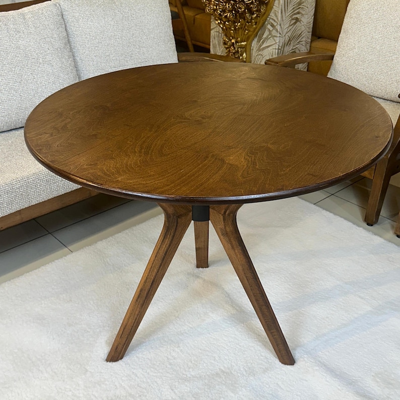 May include: A round wooden table with a dark brown finish and a visible wood grain pattern. The table features three angled legs that meet at a central point beneath the circular top, providing a stable base for the furniture piece.