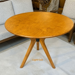May include: A round light oak table with a smooth, circular wooden top and three angled legs that meet at a central black support piece. The wood grain is visible across the surface, and the text Light Oak is printed near the base.
