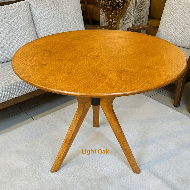 May include: A round light oak table with a smooth, circular wooden top and three angled legs that meet at a central black support piece. The wood grain is visible across the surface, and the text Light Oak is printed near the base.