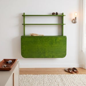 May include: A wall-mounted green wooden desk featuring a fold-down surface and two open shelves above. The desk has a visible wood grain finish and is mounted against a plain wall. Two small pinecones sit on the top shelf, and wheat stalks rest on the desk.