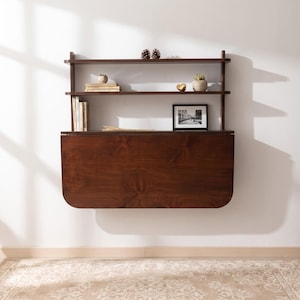 Puede incluir: Un escritorio de pared de madera marrón oscuro con una mesa abatible y dos estantes superiores integrados. Los estantes contienen varios libros, dos pequeñas plantas en macetas, dos piñas, un pequeño objeto dorado y una foto enmarcada.