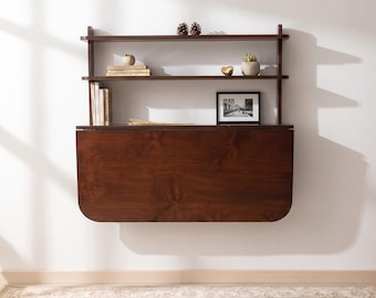 Könnte beinhalten: Ein dunkelbrauner Wandklapptisch aus Holz mit einer herunterklappbaren Tischplatte und zwei integrierten oberen Regalböden. Die Regale enthalten mehrere Bücher, zwei kleine Topfpflanzen, zwei Tannenzapfen, ein goldenes Objekt und ein gerahmtes Foto.
