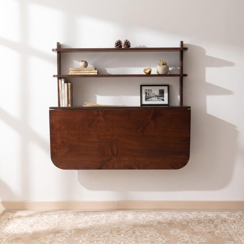 May include: A dark brown wooden wall-mounted desk featuring a fold-down tabletop and two integrated upper shelves. The shelves hold several books, two small potted plants, two pinecones, a small gold-toned object, and a framed black and white photo.
