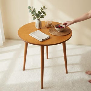 Op de afbeelding: Een ronde houten tafel met drie poten, met een vaas met groen, een kopje, een kom met bessen en een gevouwen servet. De tafel heeft een diameter van ongeveer 76 cm en staat op een wit tapijt.
