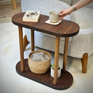 Puede incluir: Una mesa auxiliar de madera marrón oscuro con tablero y base ovalados, sostenida por cuatro patas cilíndricas. La superficie superior sostiene un libro con gafas de montura negra y una taza de cerámica sobre un posavasos. Una cesta y una planta.