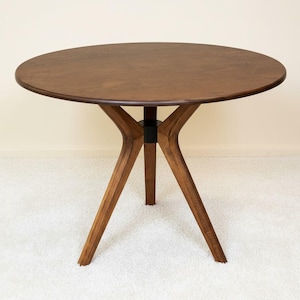 May include: A round dining table crafted from dark brown wood with a smooth, circular tabletop. The table features three angled, tapered legs that converge at a central black cylindrical support piece, creating a stable and balanced structure.