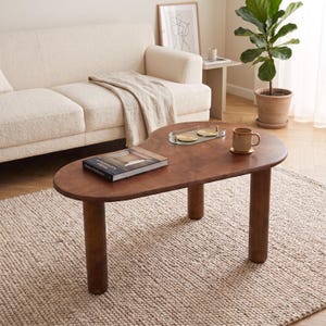 Peut inclure: Une table basse en forme de rein en bois brun foncé avec trois pieds cylindriques épais. Le plateau supporte un livre rectangulaire intitulé Modern Architecture, un plateau en verre transparent avec deux dessous de verre dorés et une tasse.