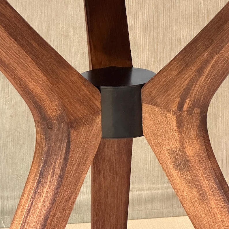 May include: A furniture base featuring four angled, medium-brown wooden legs that converge at a central, cylindrical black metal hub. The wood grain is visible throughout the legs, which extend outward from the dark, solid circular connector.