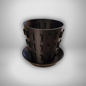 May include: A black, cylindrical planter with a matching saucer. The planter has a series of rectangular cutouts around its exterior. The design is modern and minimalist, suitable for indoor or outdoor use. The planter is made of a dark, possibly plastic material.