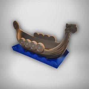 May include: A brown Viking ship model featuring a carved dragon head prow and a high curved stern. The ship has circular shields mounted along the sides and is positioned on a blue base with a stylized wave pattern representing the ocean.