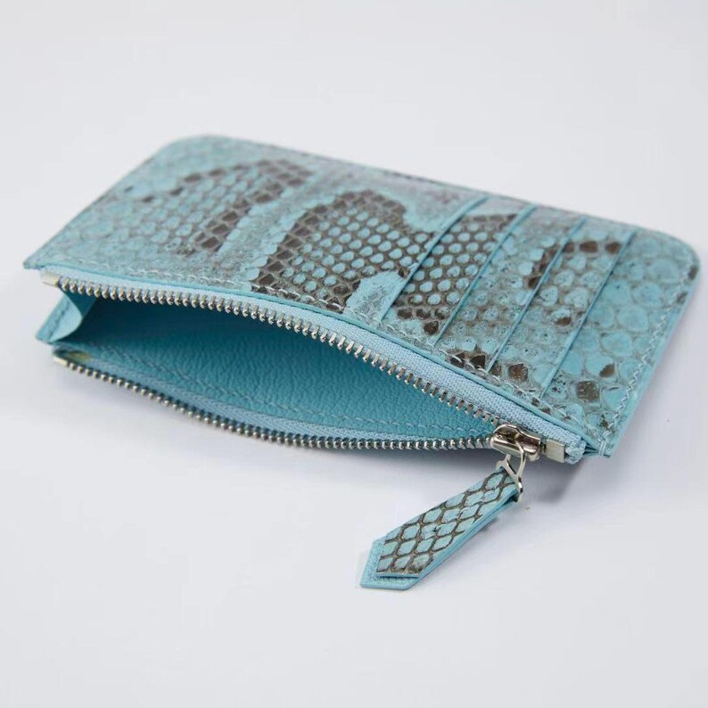 Puede incluir: Un tarjetero turquesa y gris con estampado de piel de serpiente y cremallera plateada. El interior es de color turquesa liso. El tarjetero tiene varias ranuras para tarjetas y un compartimento con cremallera.