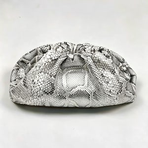 Può includere: Una pochette bianca e grigia con motivo a pelle di serpente. La borsa ha una forma arrotondata e arricciata e un aspetto morbido e strutturato. La combinazione di colori è principalmente nero, bianco e grigio.
