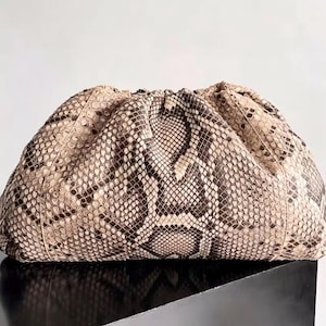May include: A structured clutch bag featuring a textured snakeskin pattern in shades of beige, cream, and dark brown. The bag has a gathered, rounded top opening with a soft, voluminous silhouette and a consistent scale print across the exterior.