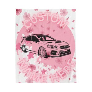 May include: A white rectangular blanket featuring a large pink circular background with scattered cherry blossom petals. A black outline of a sports sedan is centered, with the text CUSTOM NAME HERE in pink block letters above and below.