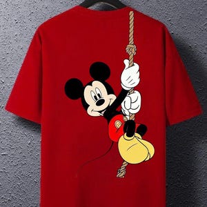 Pode incluir: Uma t-shirt vermelha de manga curta com uma estampa gráfica do Mickey Mouse pendurado numa corda vertical. A personagem é representada com orelhas pretas, rosto creme, luvas brancas, calções vermelhos e sapatos grandes amarelos.