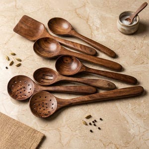 May include: A collection of dark brown wooden kitchen utensils including a flat spatula, several solid spoons, a slotted spoon, and two perforated strainers, accompanied by a small ceramic bowl containing salt and a tiny wooden spoon.