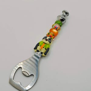 Puede incluir: Un abridor de botellas decorativo con una cabeza de acero inoxidable y un mango de cuentas. El mango presenta una figura de perezoso de dibujos animados y cuentas de colores en naranja, verde y amarillo. El abridor tiene un acabado metálico plateado.