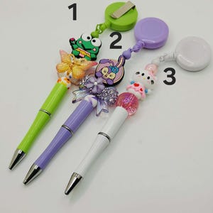 May include: Three decorative pens with retractable badge reels. The pens are green, purple, and white, each with a different charm: a frog, a bunny, and a cat. The pens have silver tips and are on a white surface.